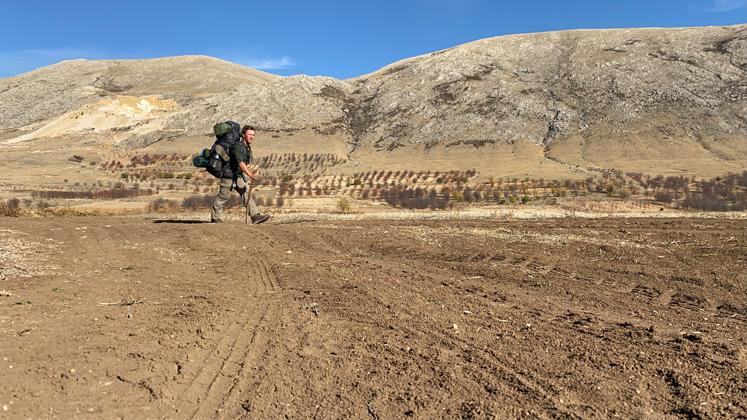 Timothy Chattell walks across a flat barren landscape with hills in the background and a deep blue sky above. He carries a large heavy backpack