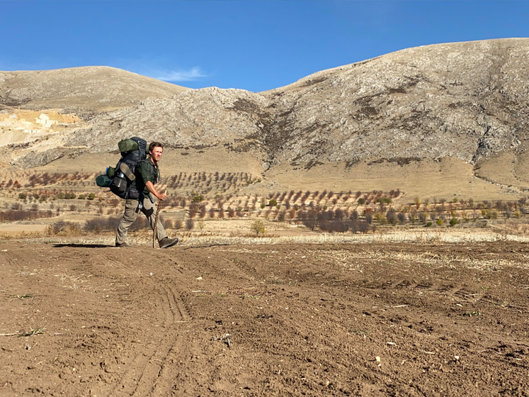 Timothy Chatell walks on flat sandy land with hills in the background and a deep blue sky. He is carrying heavy backpacks.