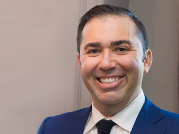 Brendan Gellman smiles at the camera. He is wearing a blue suit and a black tie