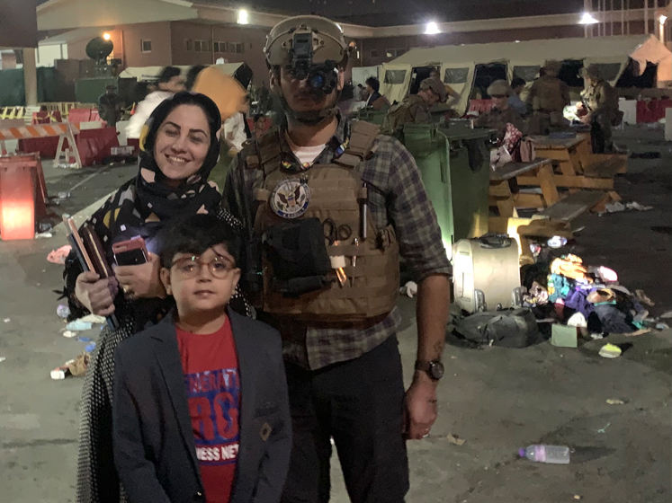 Samuel Aronson stands next to a Afghan woman and boy. The woman is smiling. Sam is wearing a security vest and helmet apparatus