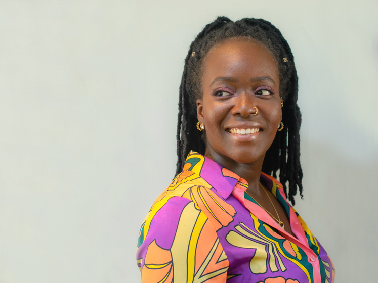 Profile image of Seyi Akiwowo showing her looking over her shoulder. She wears a colourful yellow and purple shirt and is standing against a grey background