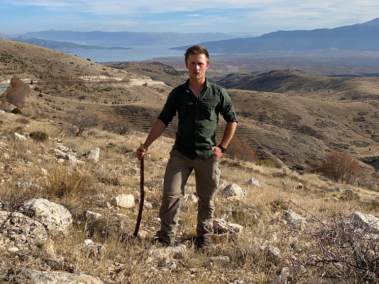 Timothy Chattell stands high on a hillside with a view of water and mountains behind him