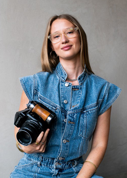 Profile image of Alice Aedy showing her wearing denim trousers and jacket and holding a camera in one arm