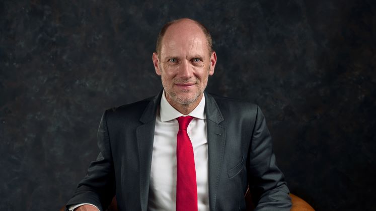 Eric Neymayer sits in a room with black walls. He is wearing a black suit, white shirt and red tie.