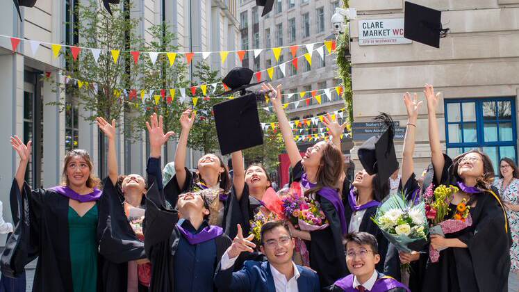 Male and female LSE graduates throw their mortarboard hats in the year while wearing graduation robes on the corner of Clare Market and Houghton Street