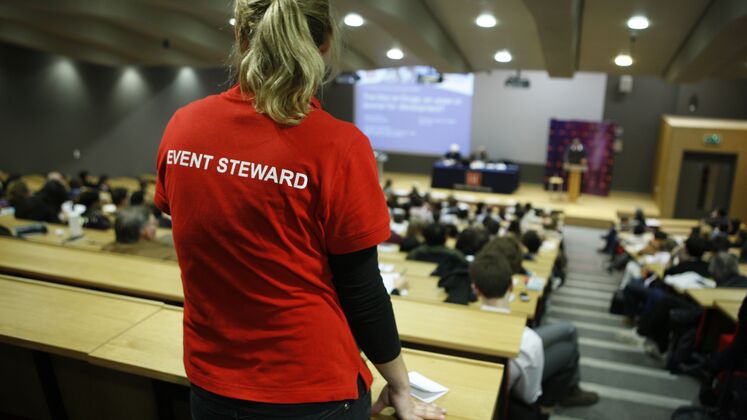 A blonde woman wearing a red event steward t-shirt stands at the top of a lecture theatre