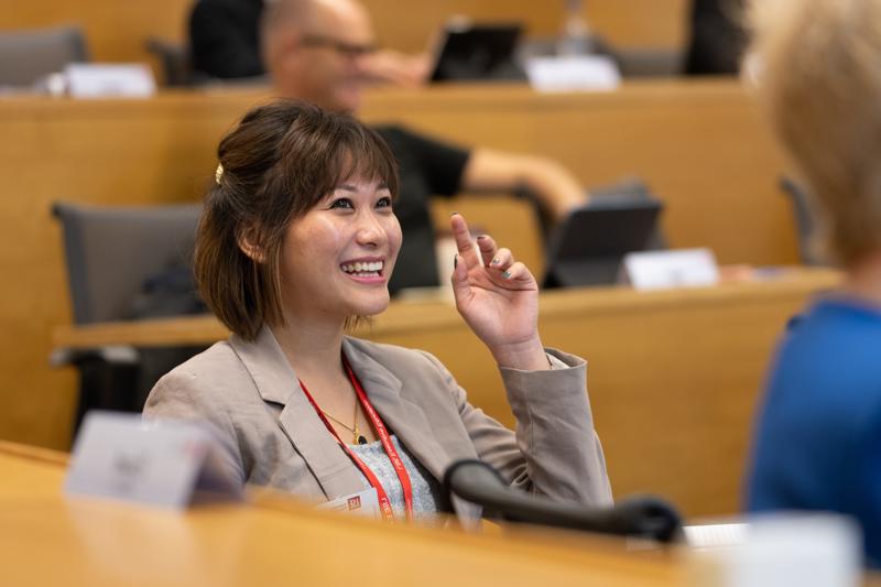 A woman sitting in a classroom smiling and putting her hand up to ask a question Sustainable Finance and Impact Investing