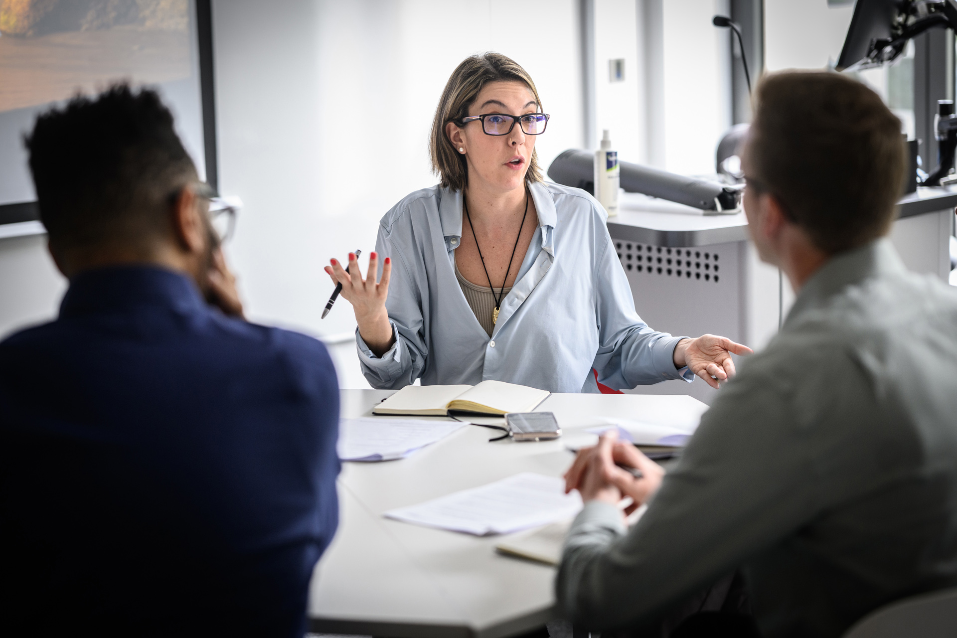 A woman with glasses speaking to 2 people from behind a desk