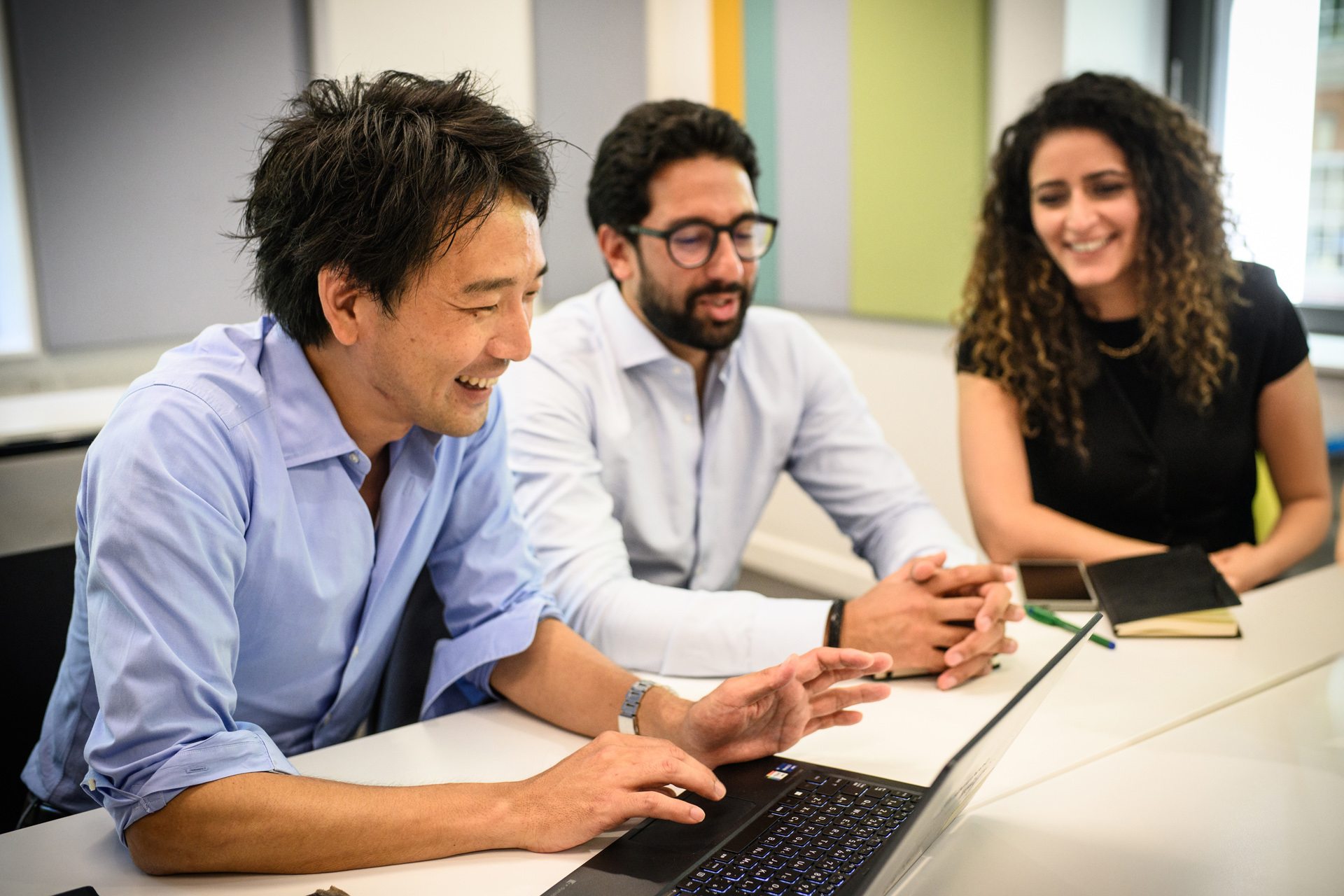 Three people sit at a desk in front of a laptop smiling
