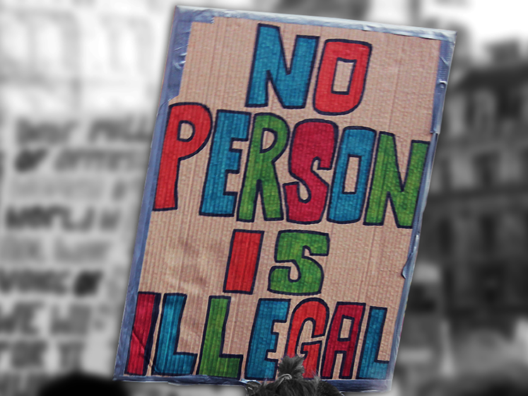 A close up of a sign at a protest which reads, 'no person is illegal'. The sign is in colour whilst the background is black and white.