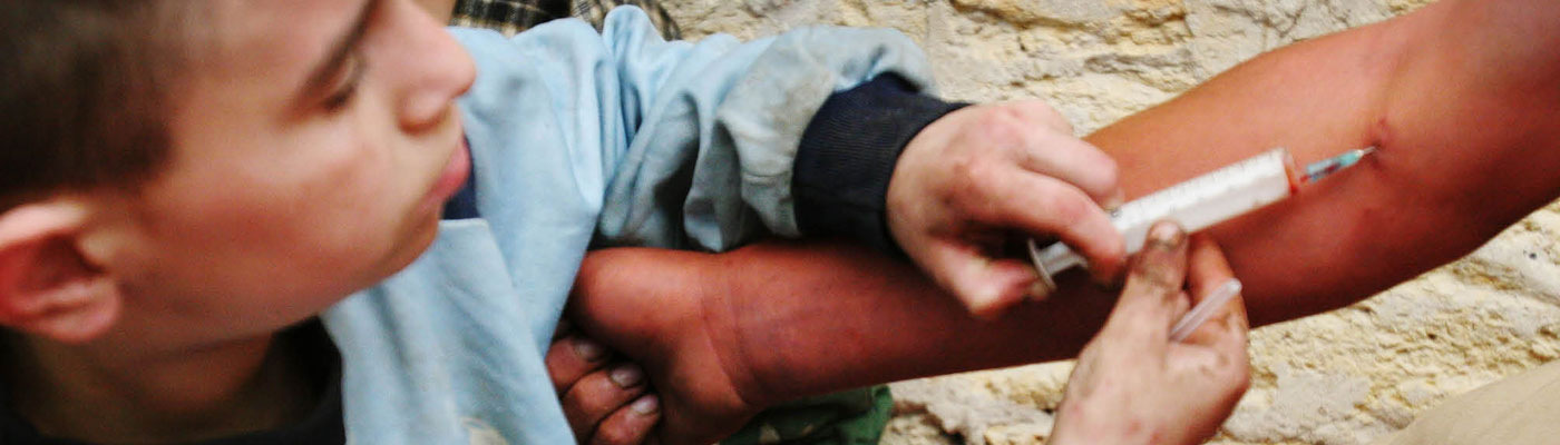 A young boy uses syringe to inject drugs into arm of an adult.