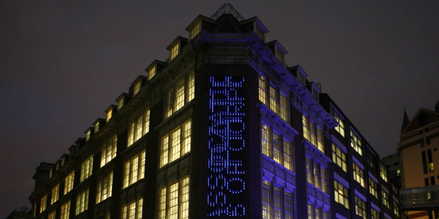 An outside view of LSE Library at night which includes the artwork Bluerain - an installation where searches from the catalogue scroll down from top to bottom on top of each other using blue LED lights.