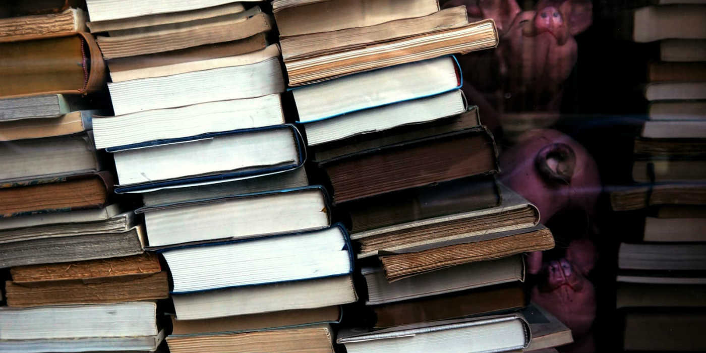 Lots of books piled up on top of each other in a few piles that are also leaning to one side because there are so many.