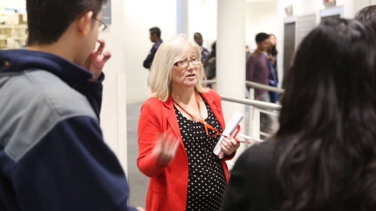 A member of Library staff wearing a lanyard is standing with a small group of students in the Library, talking to them while the students look at her and listen.