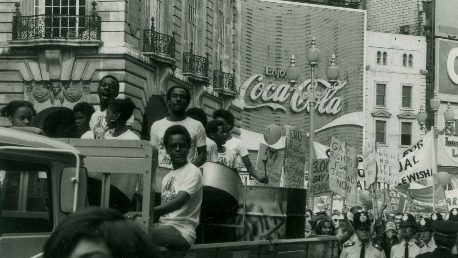 A Gay Pride procession including a group of people in a automobile and others following with placards and signs. Location appears to be Piccadilly Circus in London. There is a huge Coca-Cola sign in the background.