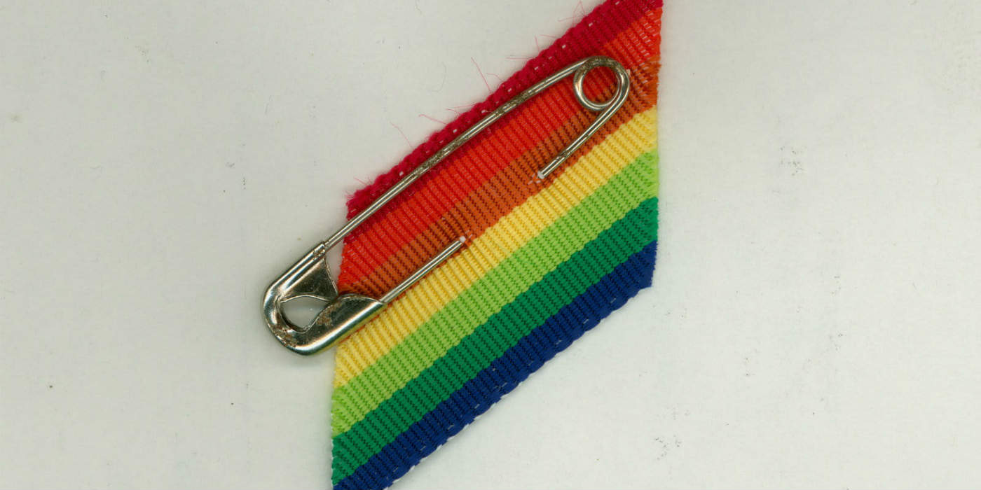 A rainbow coloured ribbon with a safety pin attached to it up against a light background.
