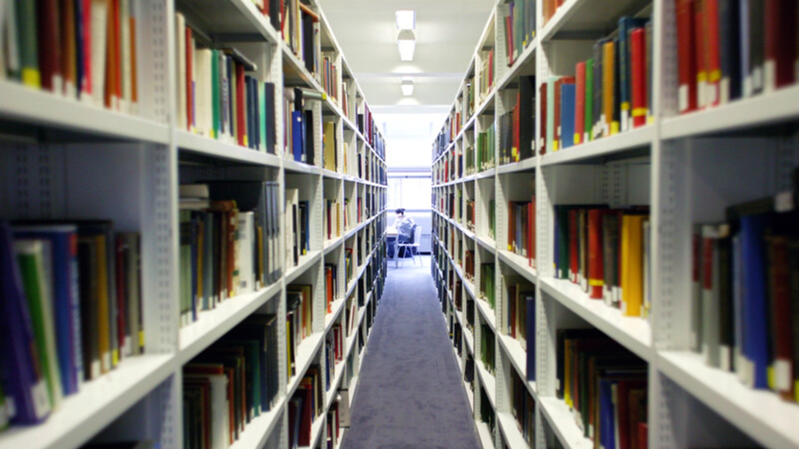 A view between two book stacks looking outward towards a study desk where a student is seated in the distance.