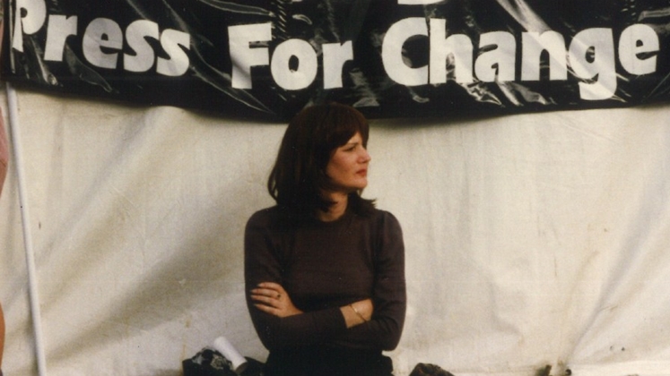 A person sat up against a tent with a sign behind them. Sign reads "Press For Change".