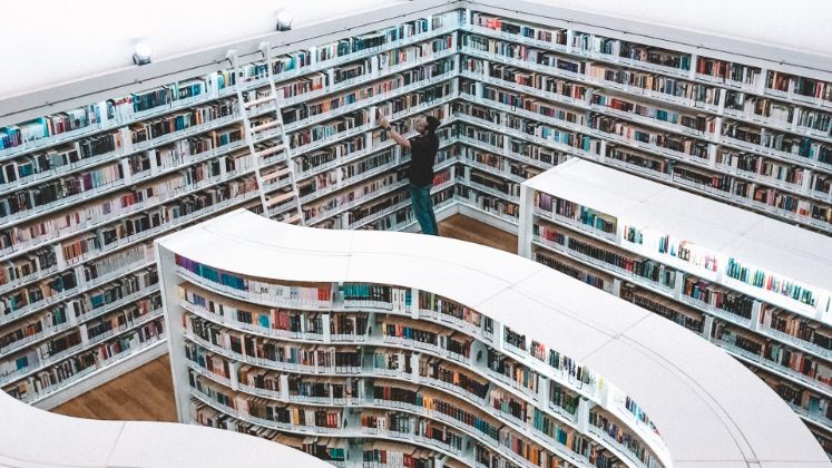 A man is standing in a library amongst several tall bookshelves full of books. He is standing close to one of the bookshelves, carefully looking up at the books.