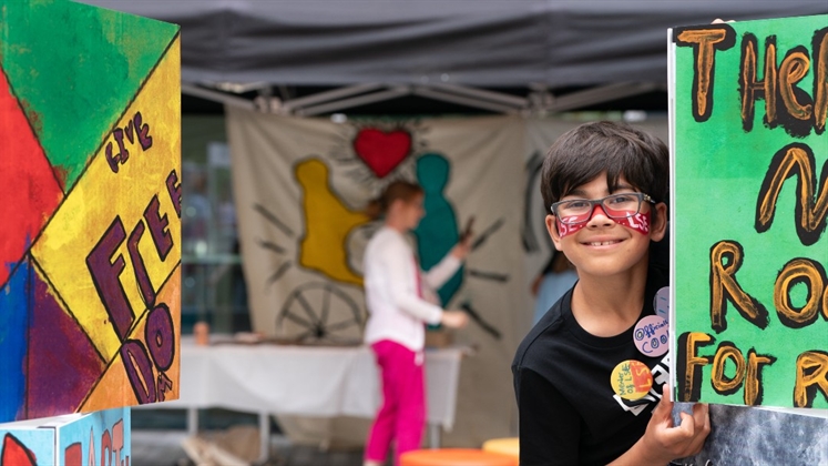 A child smiling and peering around a colourful piece of artwork for the camera.