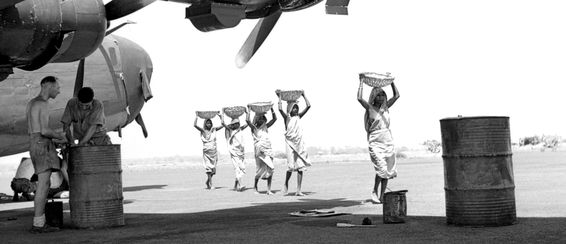 A group of women wearing saris walking along the tarmac of an airplane runway carrying goods on their heads