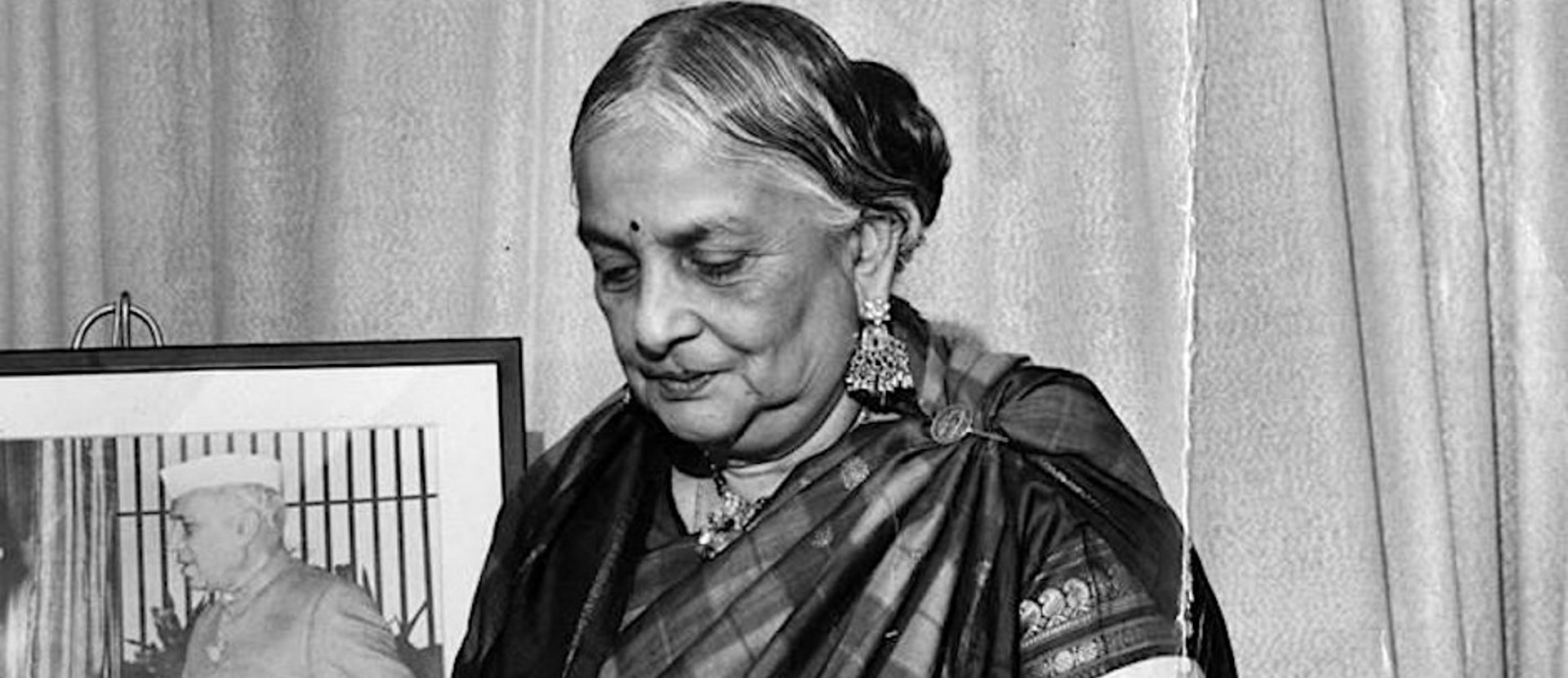 A women in Indian dress looking downwards. There is a picture of a person in a frame behind her.