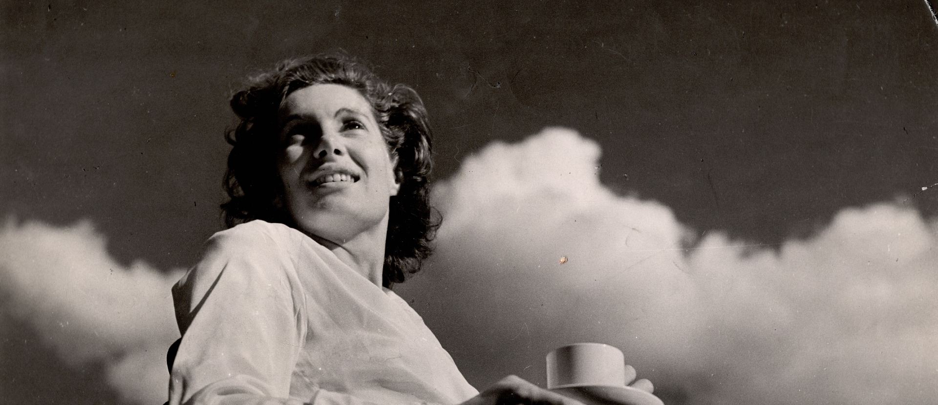 Jill Craigie photographed from below as she is seated with a cup and saucer. She is looking up into the sky and there are clouds behind her.