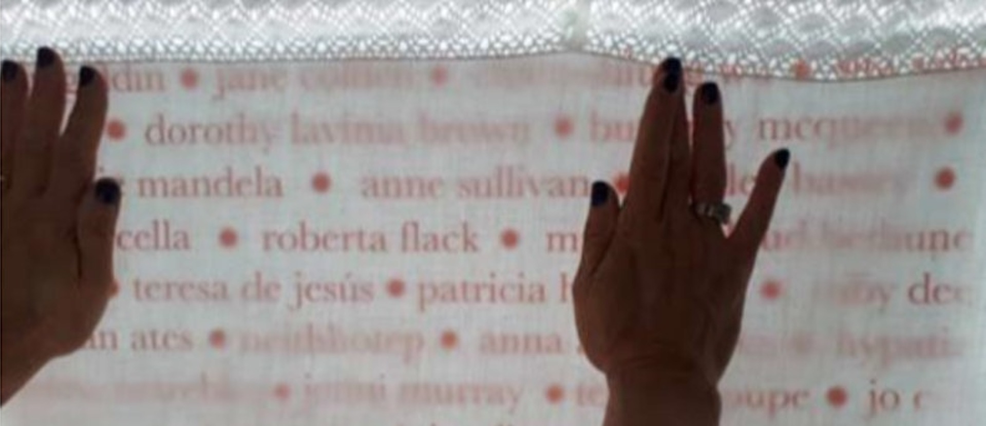A piece of textile with women's names written on. It is being held up against a light by two hands that are visible.