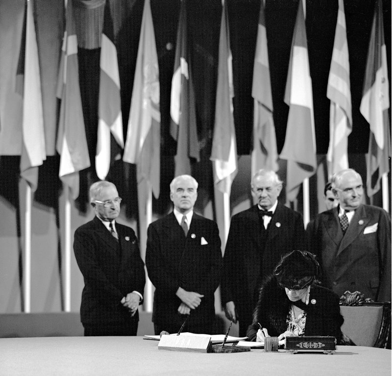 A woman sat at a desk writing. Behind her are a line of suited men and an array of flags