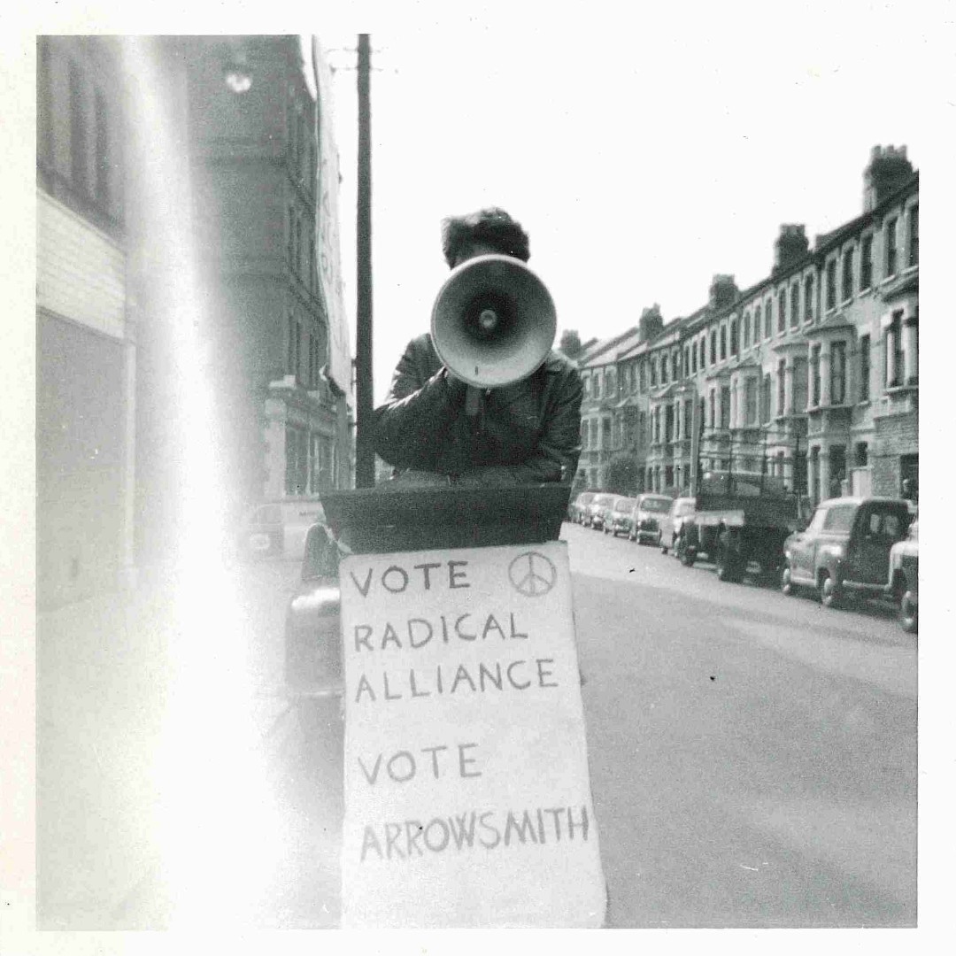 A person stood in the street, their face obscured by a megaphone. Beneath them is a sign with a CND symbol on that reads as follows "Vote Radical Alliance. Vote Arrowsmith".