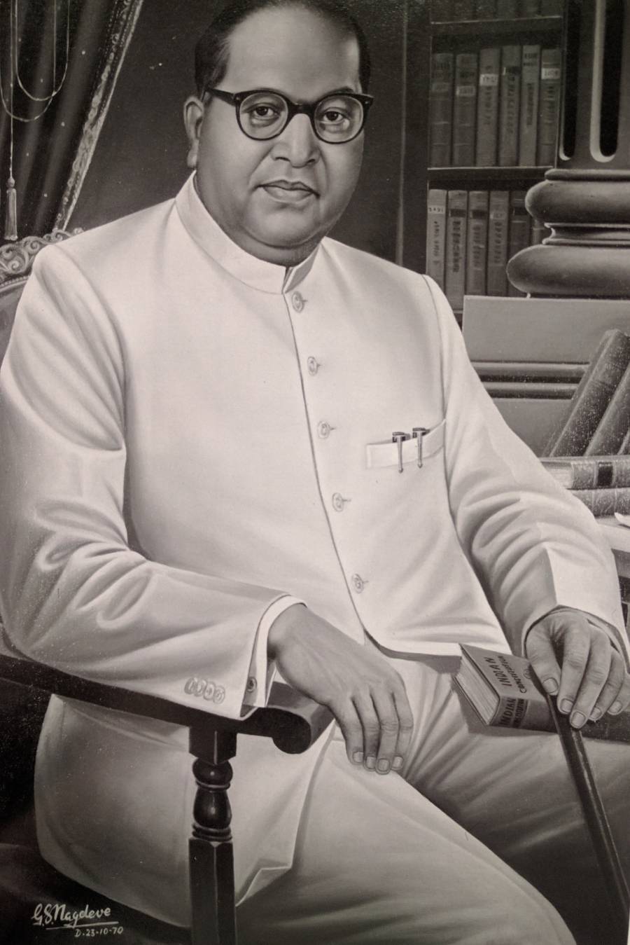 A portrait of Ambedkar sitting in a chair in front of bookshelves