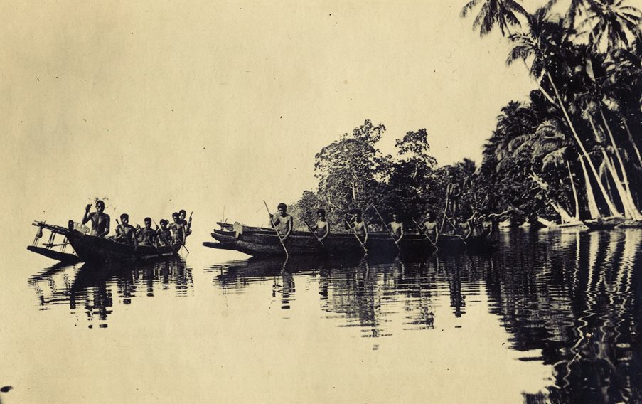 A photograph across water of two canoes being sailed by Trobriand islanders. There are trees in the background.