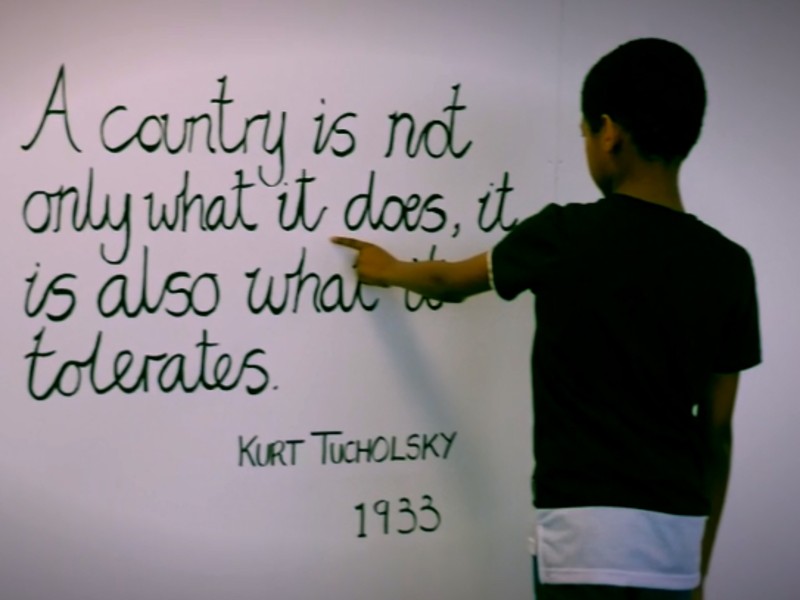 A child is pointing at a quote: "A country is not only what it does, it is also what it tolerates", Kurt Tucholsky 1933.