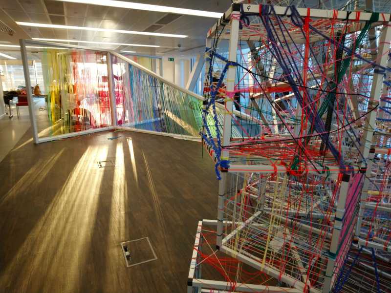 Artworks made from different coloured string woven on to cube shaped frames clustered together. In the background is a large frame with threads woven vertically in a rainbow effect.