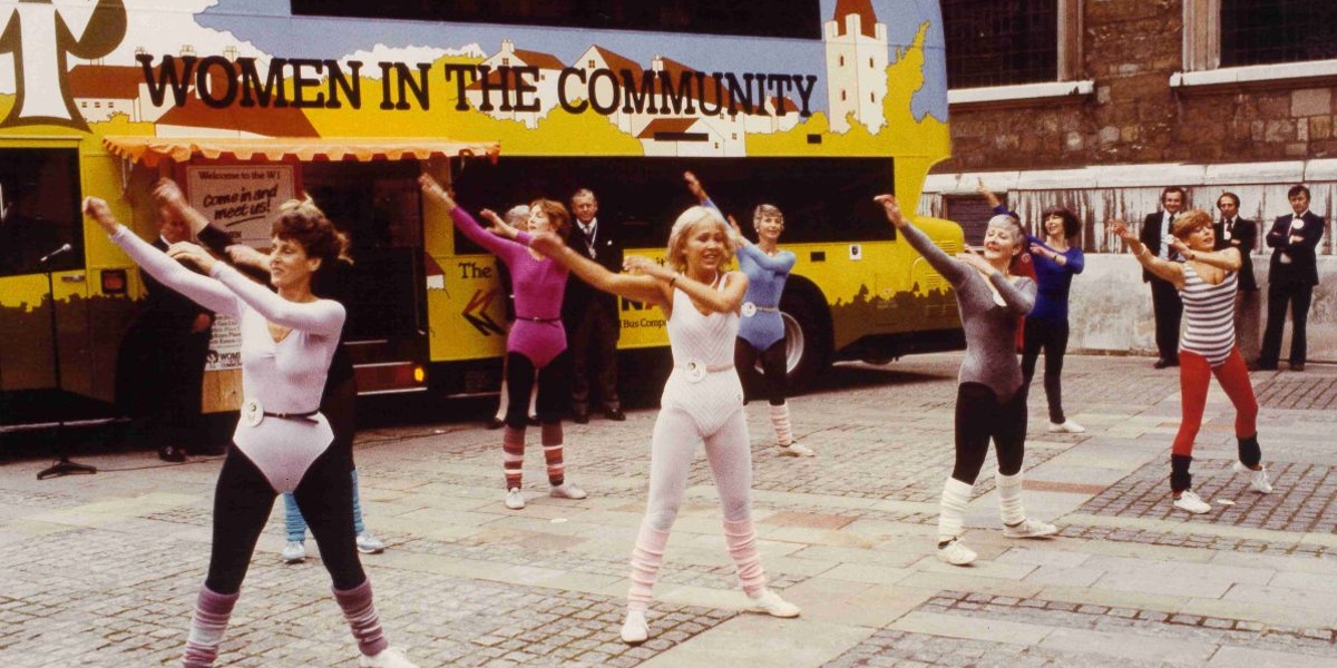 A group of people doing aerobics in the street. There is a bus behind them with Women in the Community written on it.