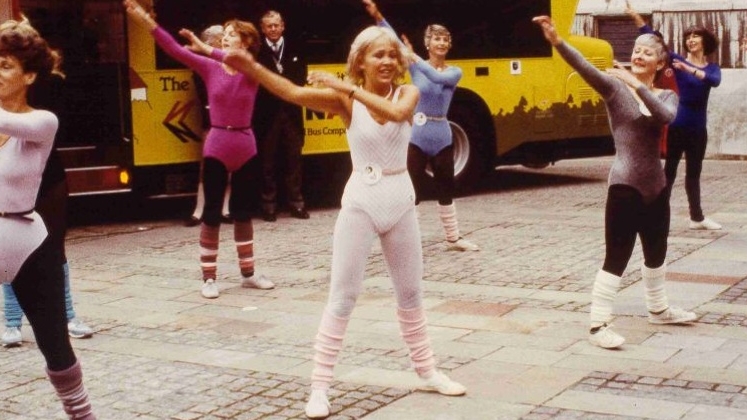 A group of people doing aerobics in the street. There is a bus behind them with Women in the Community written on it.