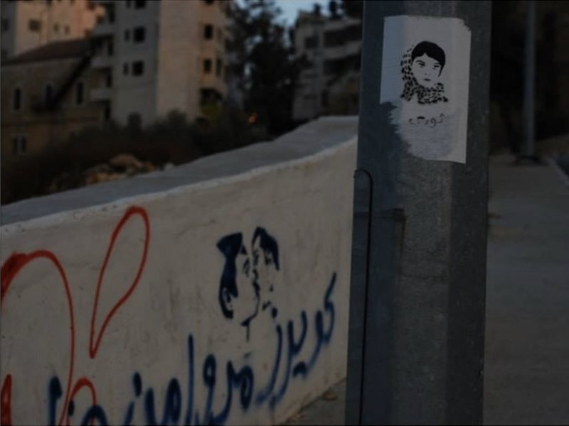 Street art of two men kissing and a poster of a woman in a hijab on a road wall in Palestine.