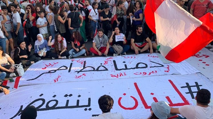 Protestors sitting around a banner saying 'Our blood is a red line #Lebanon revolts' with names of towns around the edges.