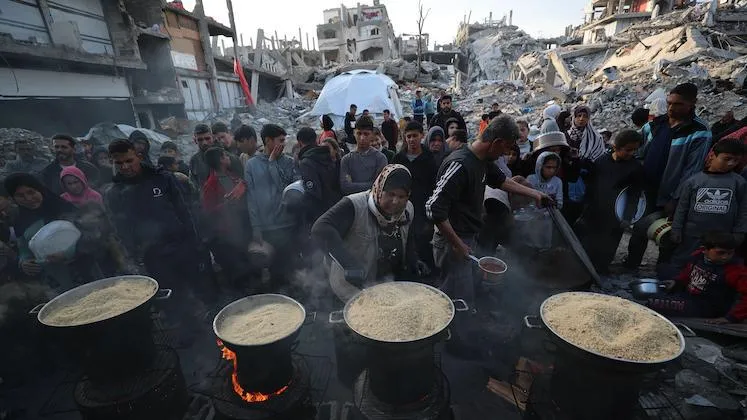 Women serving food from four large pots to a group of people, among the rubble of destroyed buildings in Gaza.