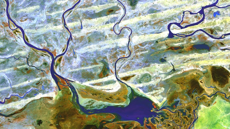 Ariel shot of a river with multiple tributaries feeding into it. The land around the river is a combination of white, green, brown and yellow. The water is deep blue.