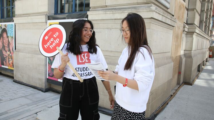 LSE open day volunteers stand on campus with a 'here to help sign'