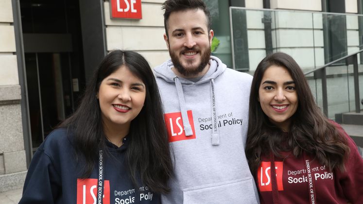Three students wearing hoodies smile at the camera while standing outside LSE's New Academic Building