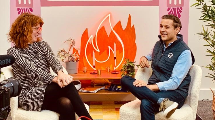 A woman and a man sit on two seats in front of a fire symbol while being filmed by a camera crew