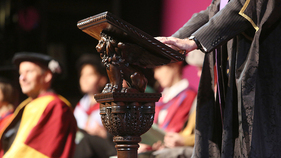 A person in academic robes stands at an old carved lectern during graduation