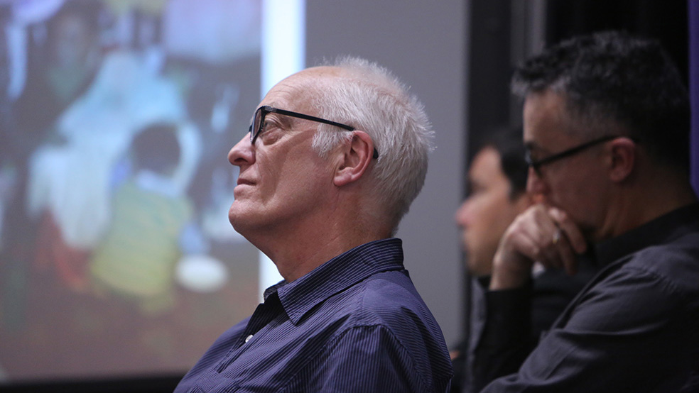 A white haired man with glasses listens to a lecture